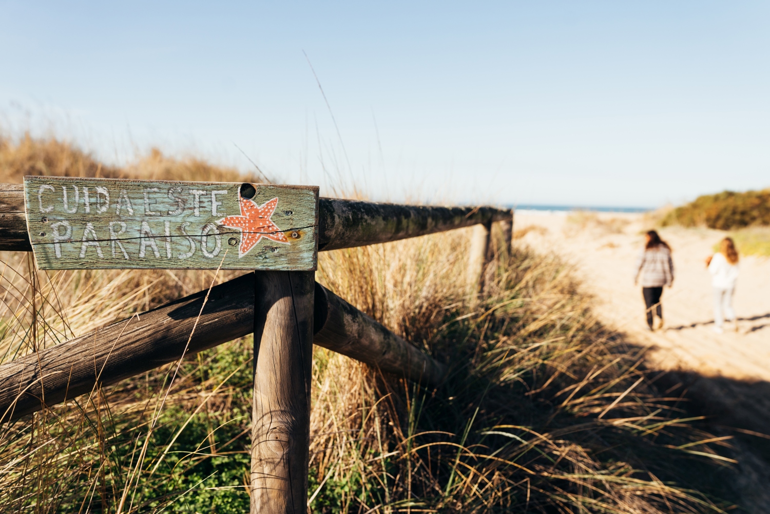 Beach sign in foreground with two people walking onto a beach in the background