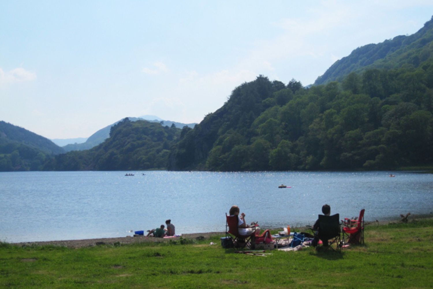 Family sat on grass beside lake with mountains and trees in background, Snowdonia