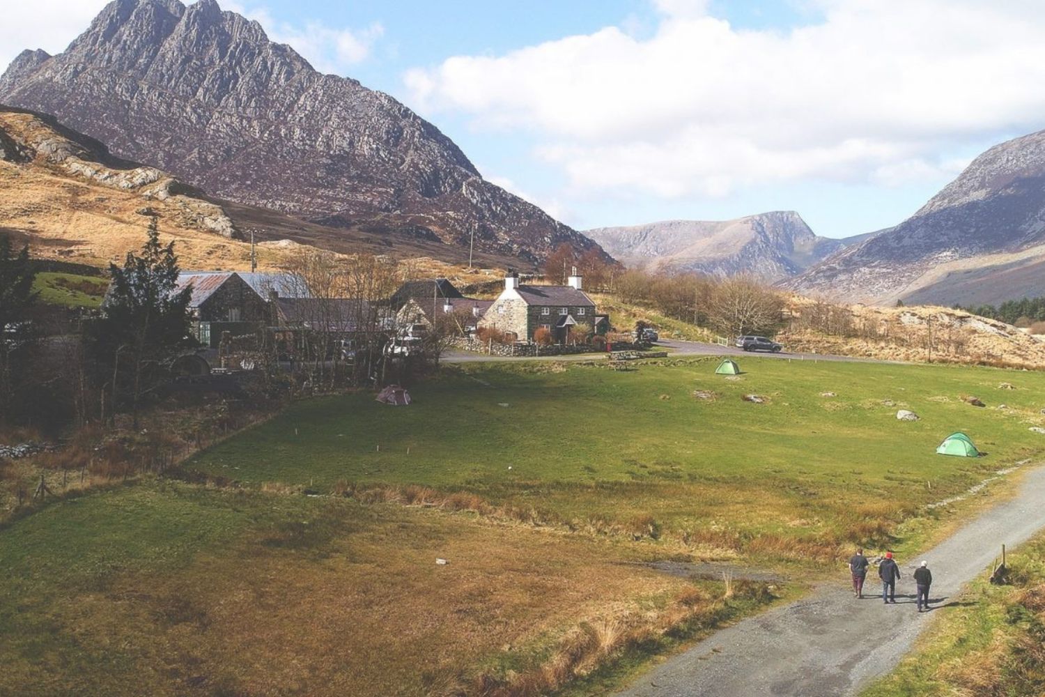 People walking along path next to farm houses and field with rocky mountains in background, Snowdonia