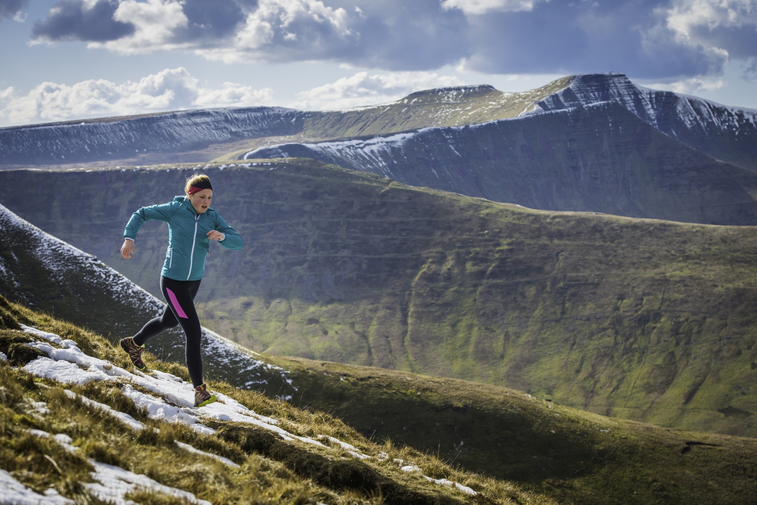 Trail runner woman running along snowy hilltop with mountains in background_Climbing Pen y Fan, Wales